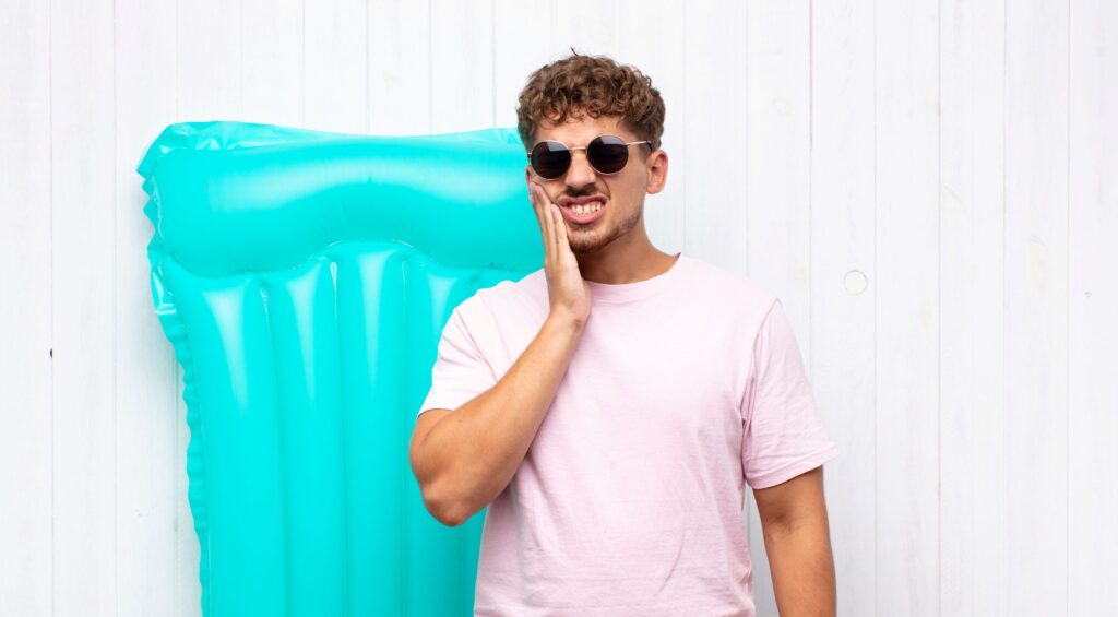 Man in pink t-shirt and sunglasses next to pool floatie holding hand to jaw in pain